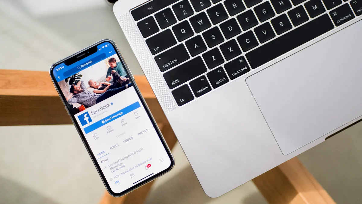 Mobile phone with the facebook website featured next to a laptop on a table.