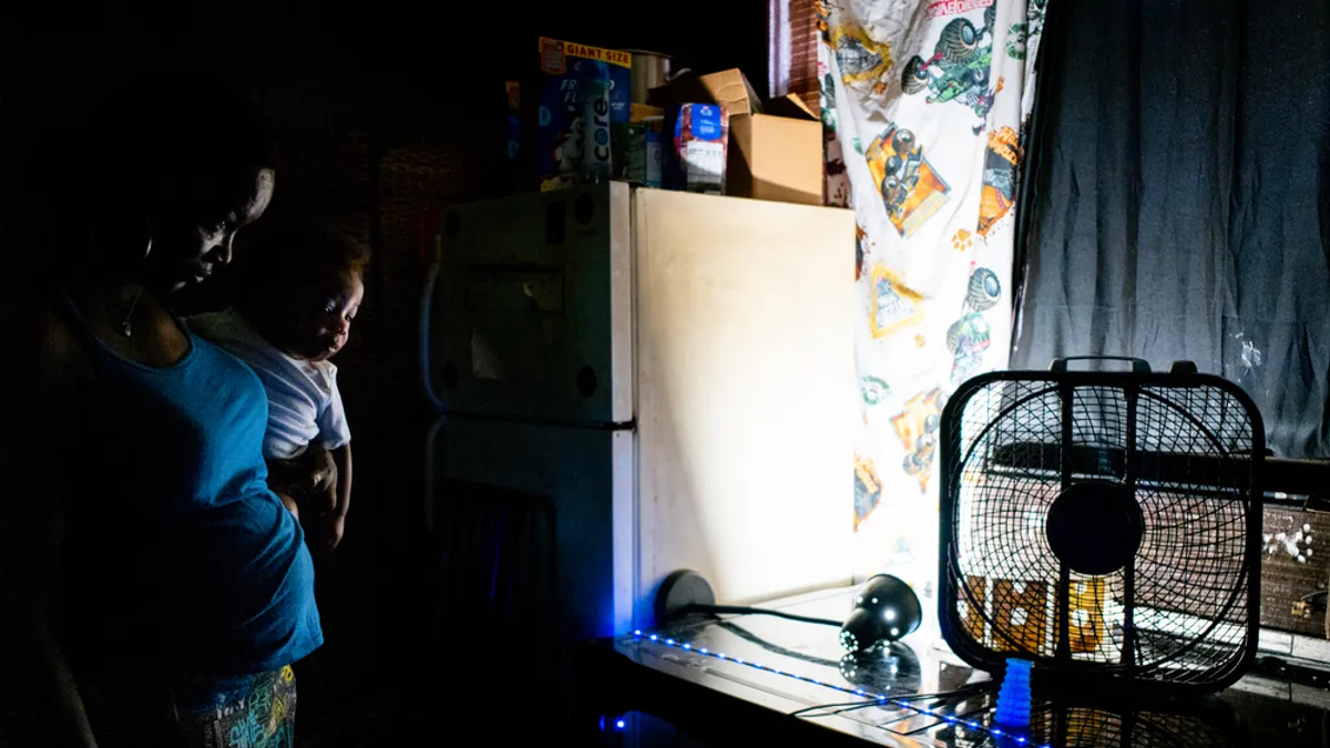 A woman holding a young child in a darkened room next to a fan.