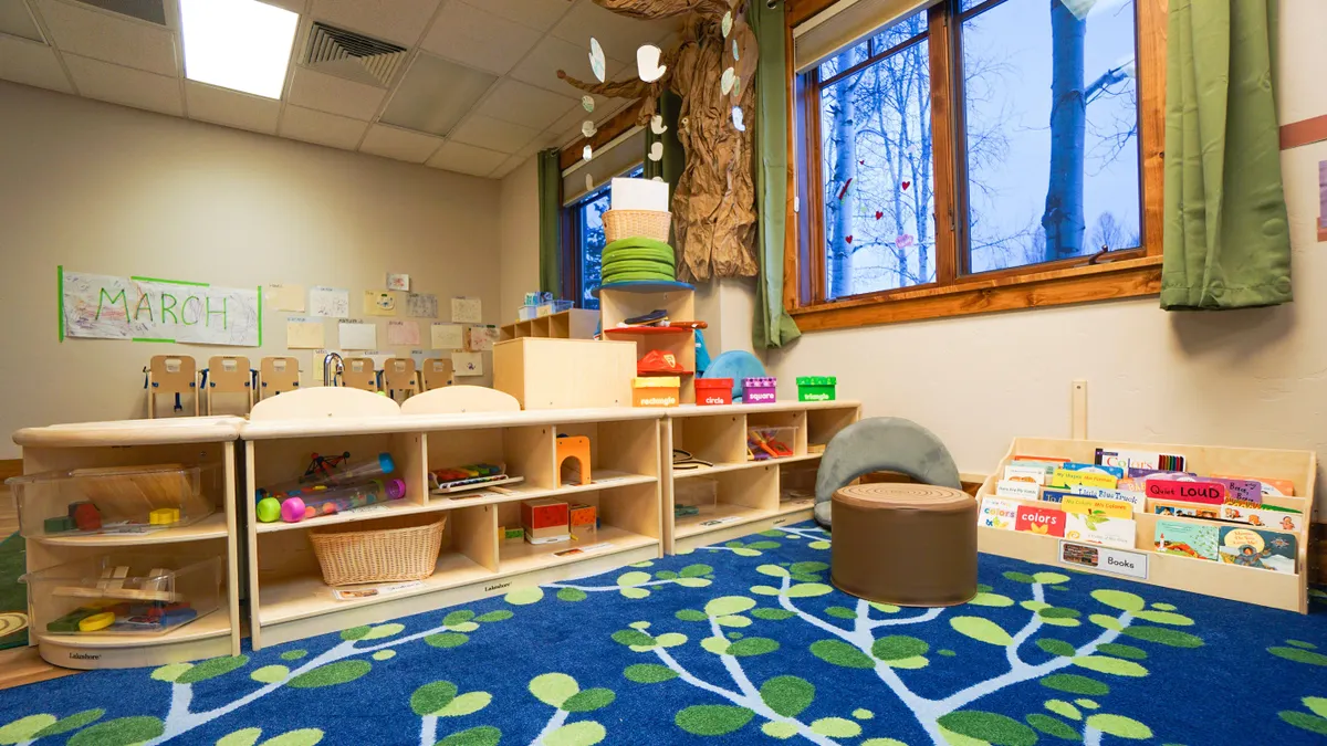 A reading and activity nook in a day care is shown, with a blue carpet, a window in the top right, and books and toys featured.