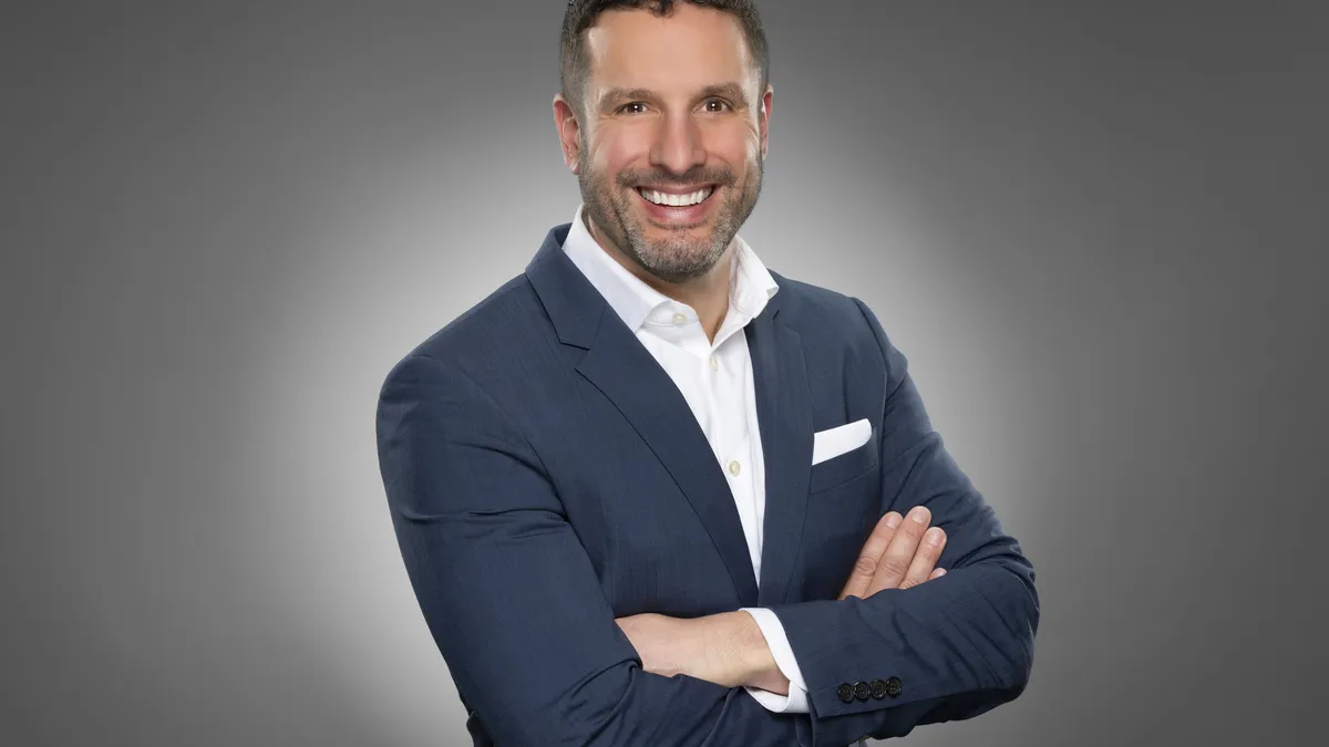 A person with short dark hair and a beard smiles with arms crossed, wearing a white shirt and blue suit coat, no tie.