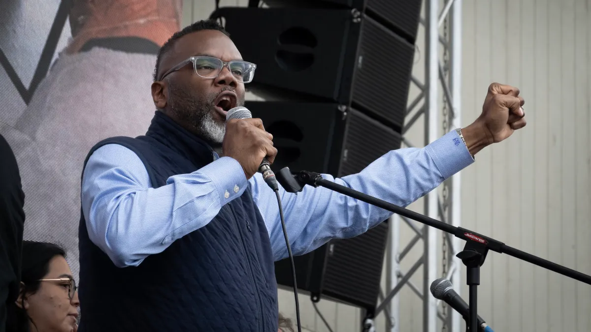 A person standing in front ot a mural speaks into a microphone and raises his fist.
