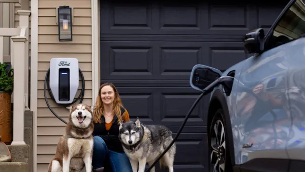 A person kneels with two large dogs in front of a garage with a Ford residential EV charger mounted on a wall.