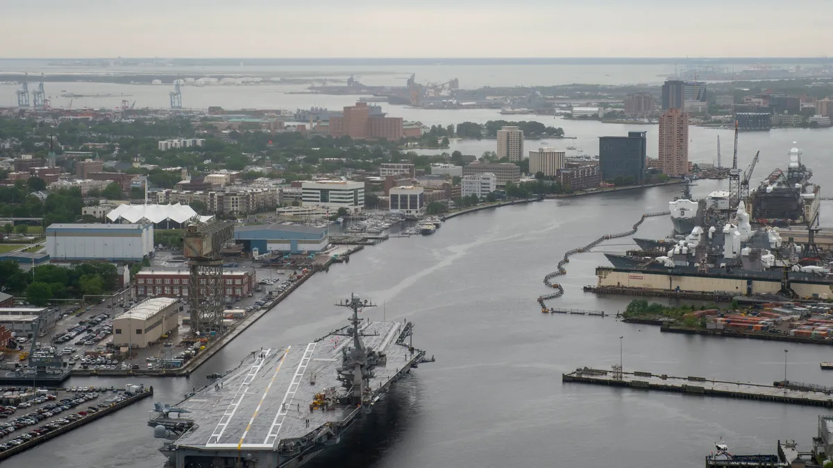 An aerial view of an aircraft carrier navigating the Elizabeth River as it departs Norfolk Naval Shipyard in Virginia