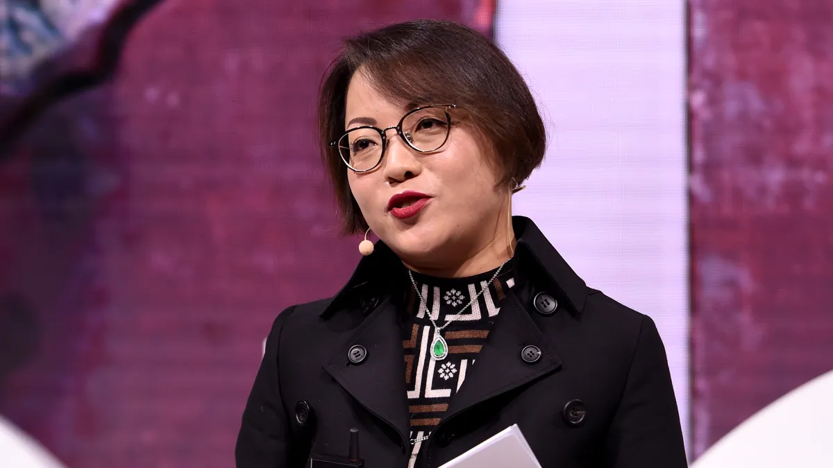 A person stands on stage wearing a headset and speaking to the audience.