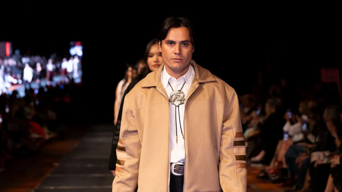 A person in a tan jacket walks in front of a line of other people on a white runway as a crowd watches from the sidelines.