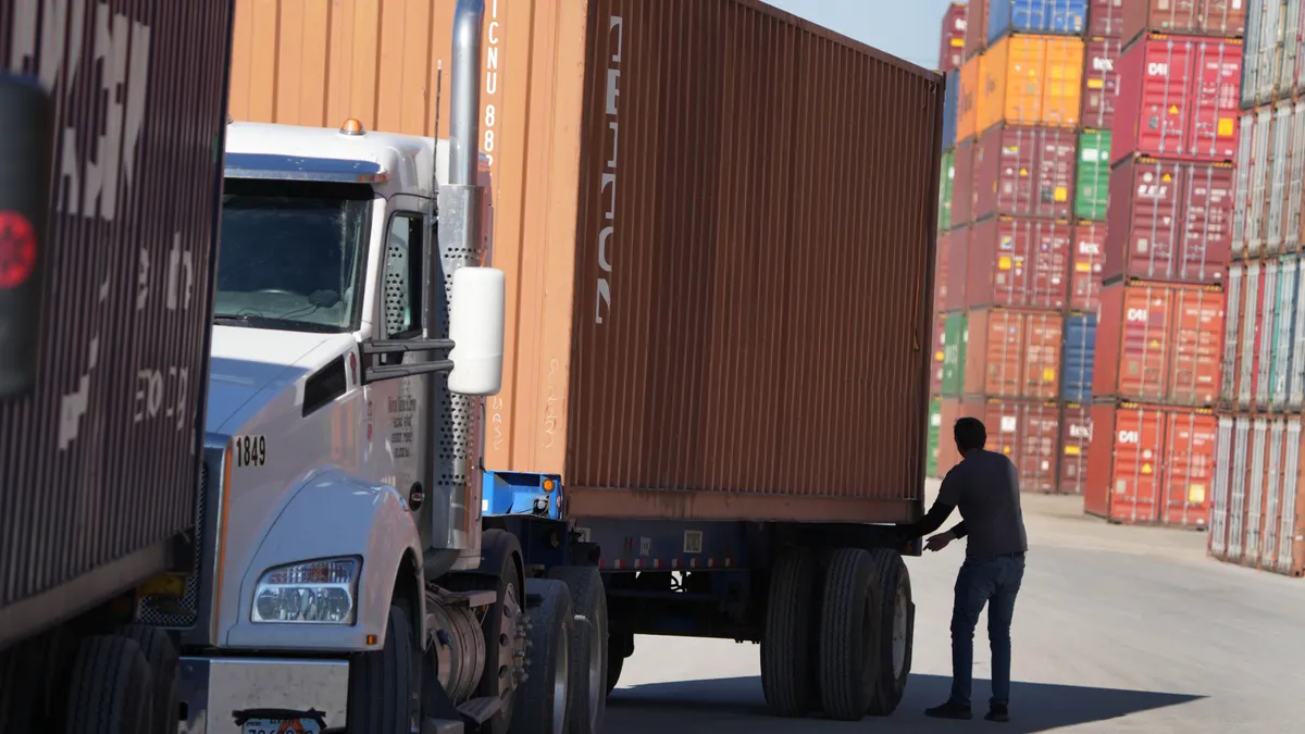 A truck driver stands behind a semitruck carrying an orange container. Stacks of shipping containers can be seen in the background.