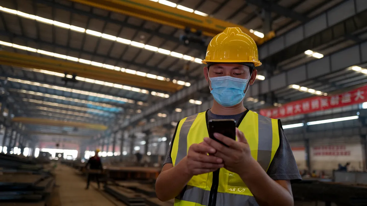 An employee wears a face mask and uses a mobile phone on the job at a construction site.