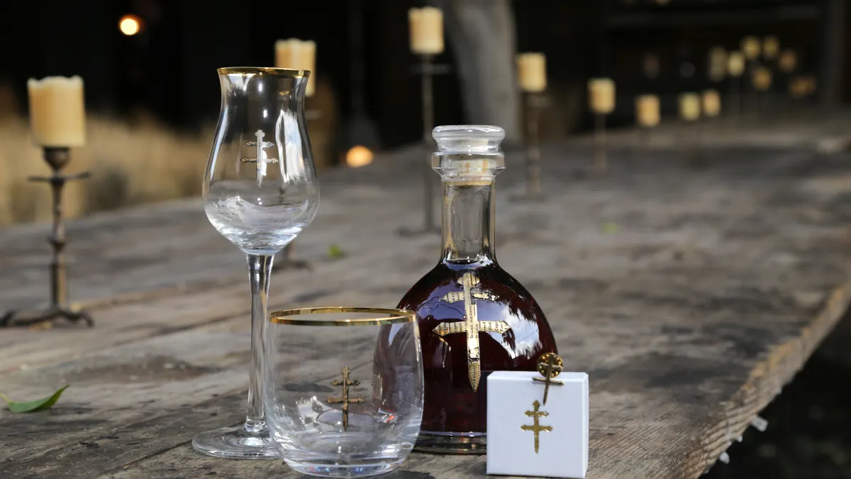 A bottle of D'Usse cognac sits on an old wooden bar next to a gold-rimmed old fashioned glass and cocktail glass, both which have the gold D'Usse cross. A small white box is next to the bottle.