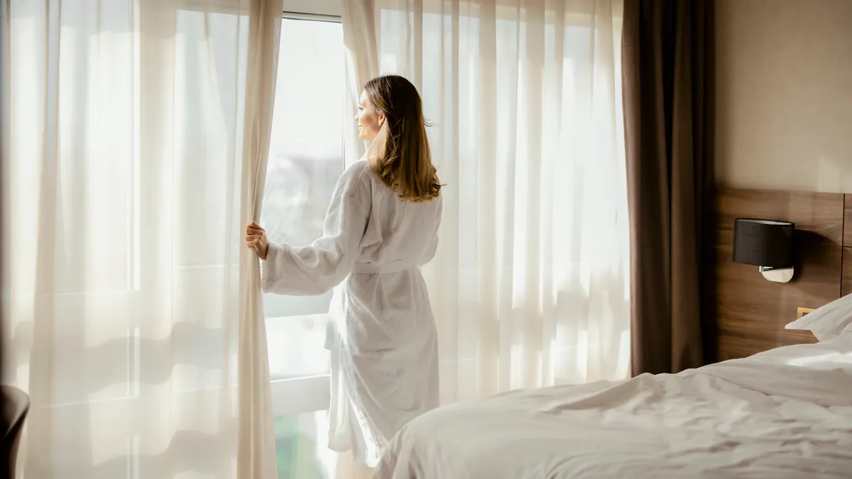 A woman looks out of a hotel room window.