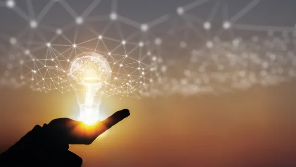 At sunset, a human hand holds a lightbulb with a transparent brain inside and a digital webbed grid emanating outward