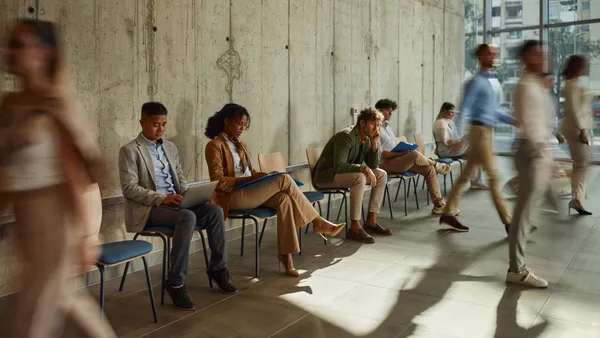 People sit in chairs against a wall while some get up to walk.