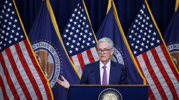 A man with silver hair in a suit gestures at a podium.