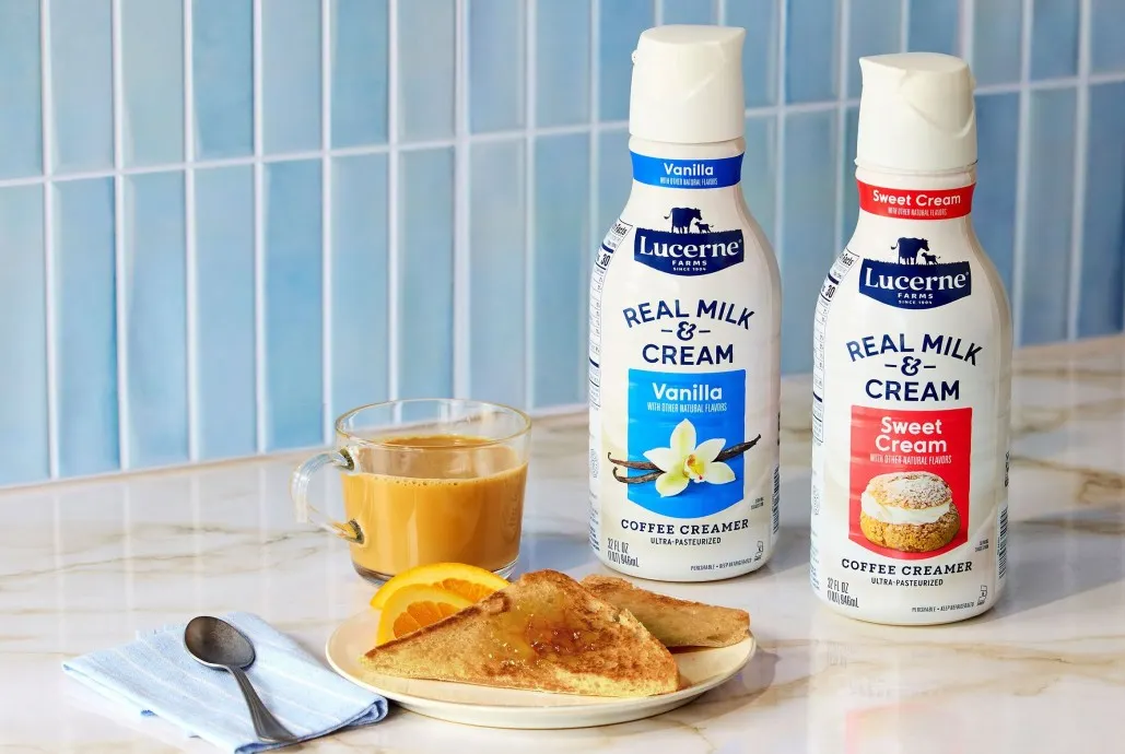 Containers of Lucerne coffee creamer displayed next to a cup of coffee, a spoon on a napkin and a plate with bread and fruit.