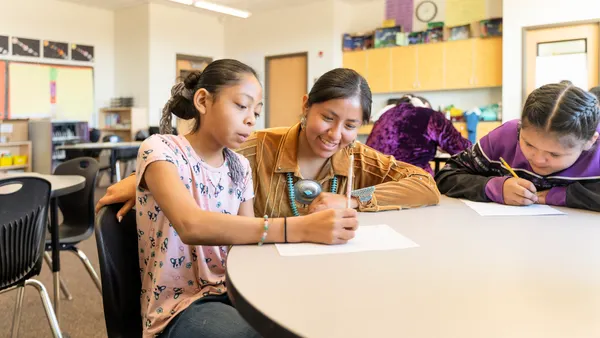 An adult is with two students at a desk in a classroom. The students are writing and reading.