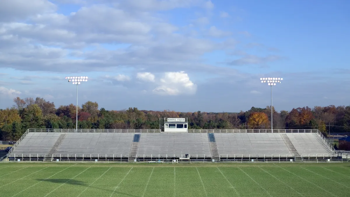 A wide camera shot of one side of the outdoor stands at a grass-covered sports field. Stadium lights are over the stands and in the background are trees and a blue sky with clouds.