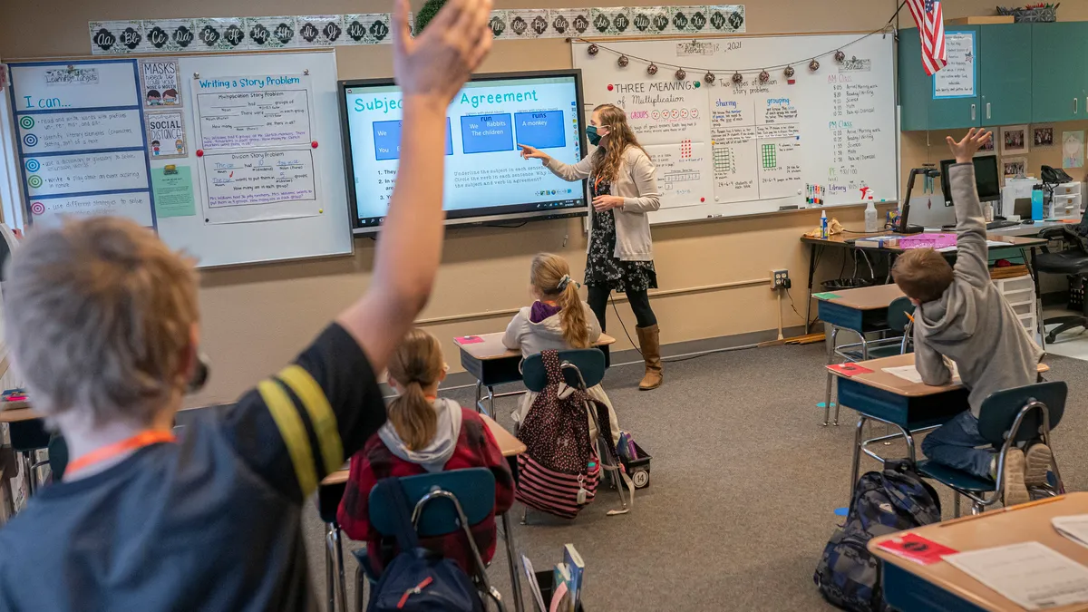 A 3rd grade boy raises his hand as a teacher explains a grammar lesson on a projection screen.