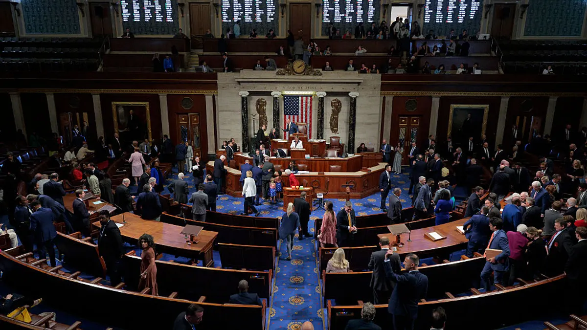 A wide-angle of the floor of the House is seen with people walking around and sitting at desks.