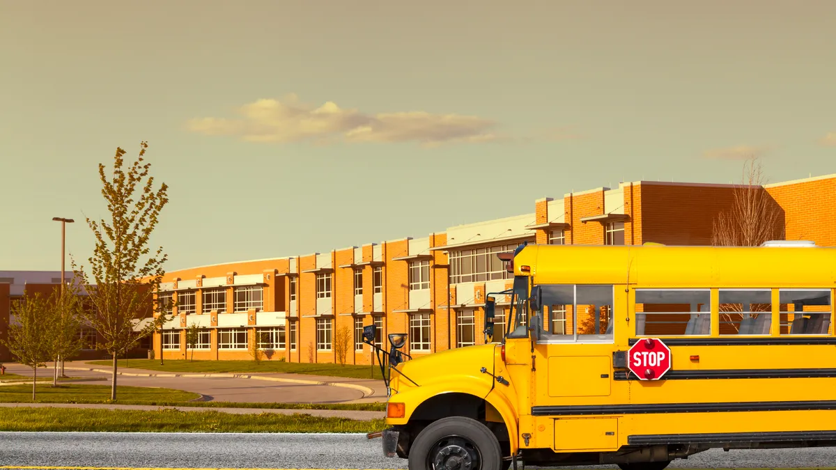 A yellow school bus is parked outside a two story level school. A light blue sky and small cloud are in the top half of the photo.