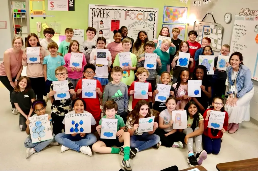 A group of elementary students in a classroom hold signs alongside their two teachers with everyone facing the camera.