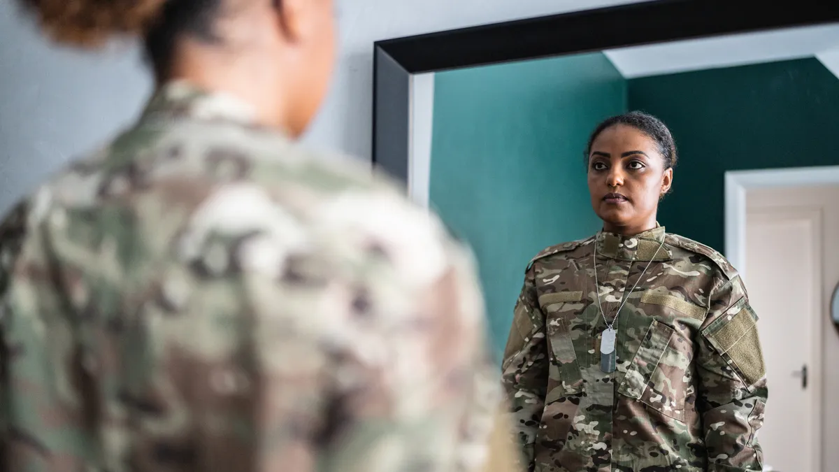 A Black woman, in military uniform, contemplates her reflection in the mirror