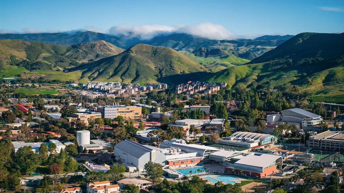 A skyshot of a large cluster of buildings in the foothills of green mountains during the daytime.