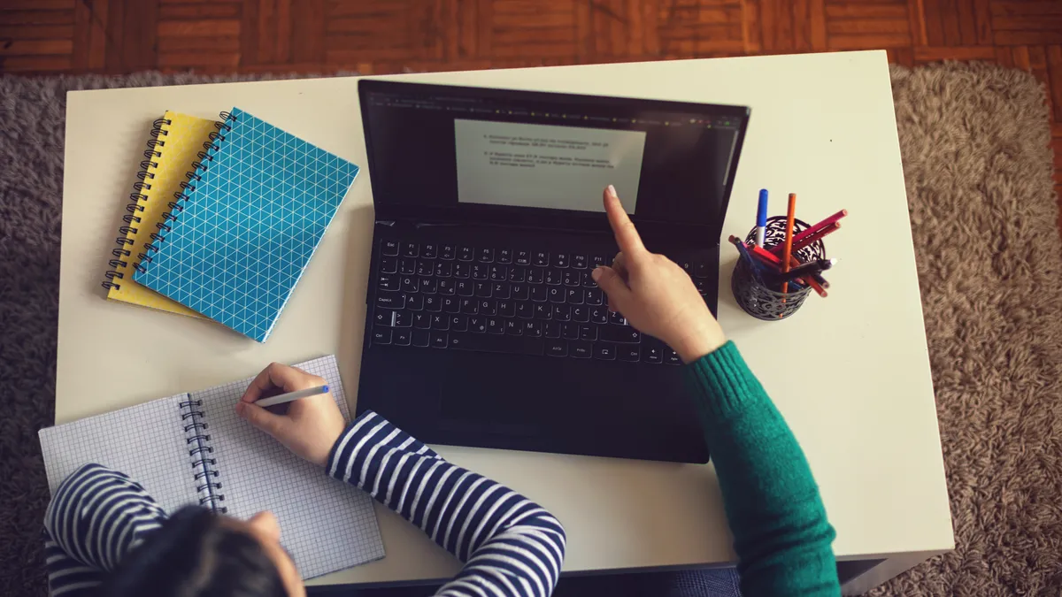 A child writes on a notepad while an adult points to a laptop screen while they are both home.