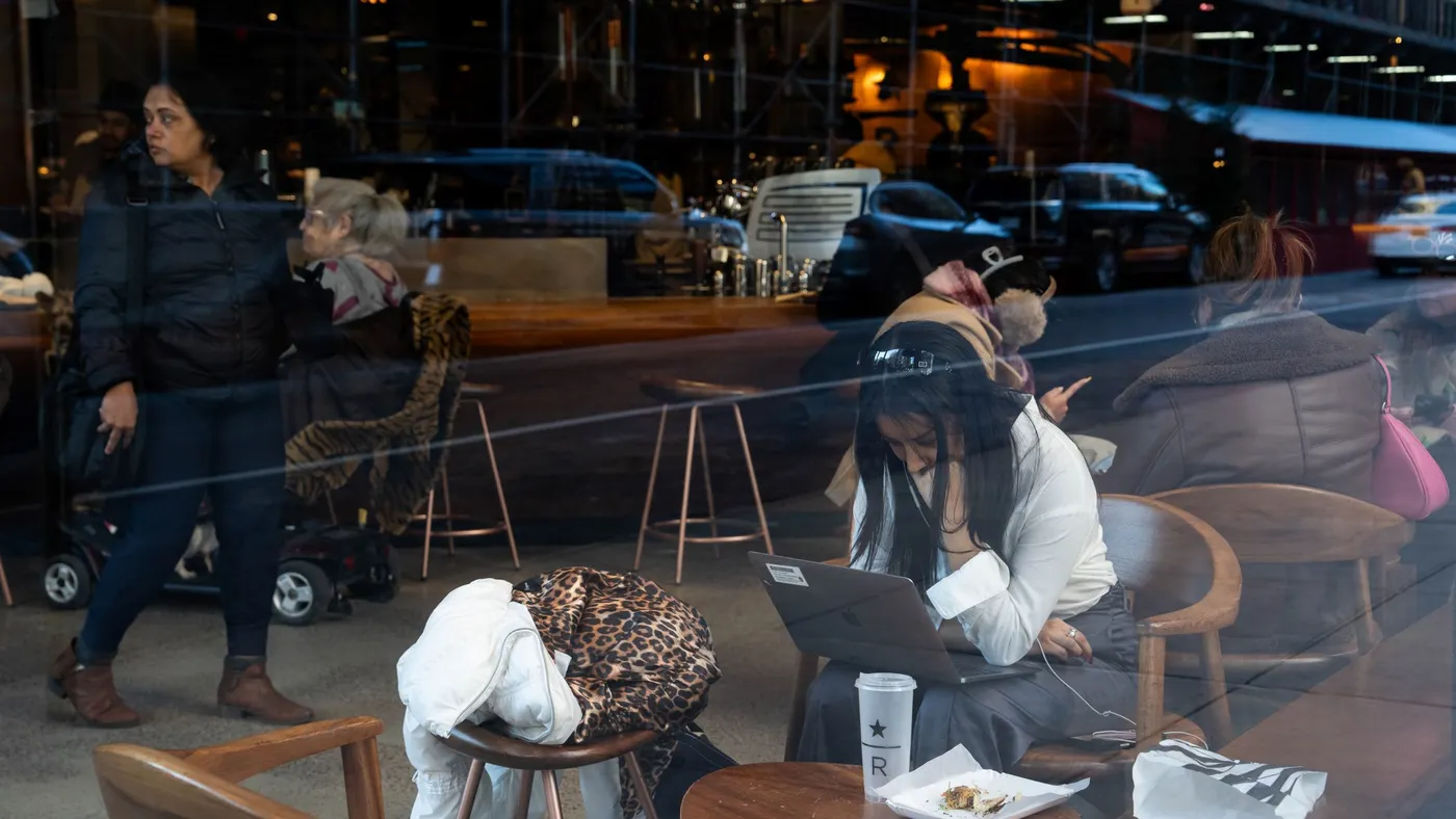 A person is seen through the window of a coffee shop, working on a laptop.