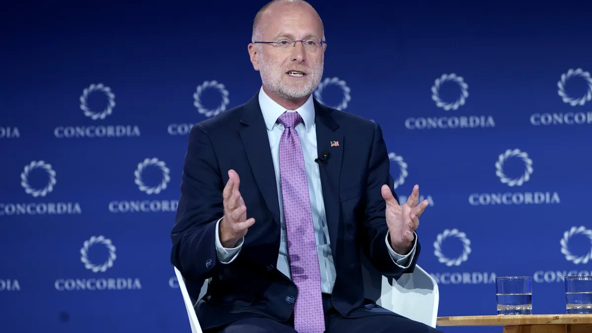 A man wearing a blue suit and purple tie gestures with both hands while sitting in a chair on a stage