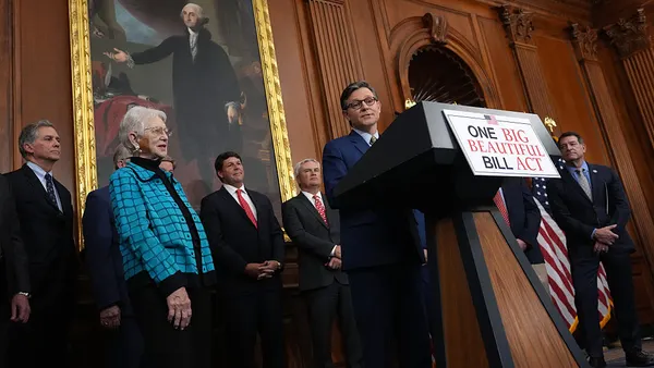 One person is standing behind a podium with a sign that reads "One Big Beautiful Bill Act." A handful of other people are standing around and behind the person and they are in a room with wood paneling.