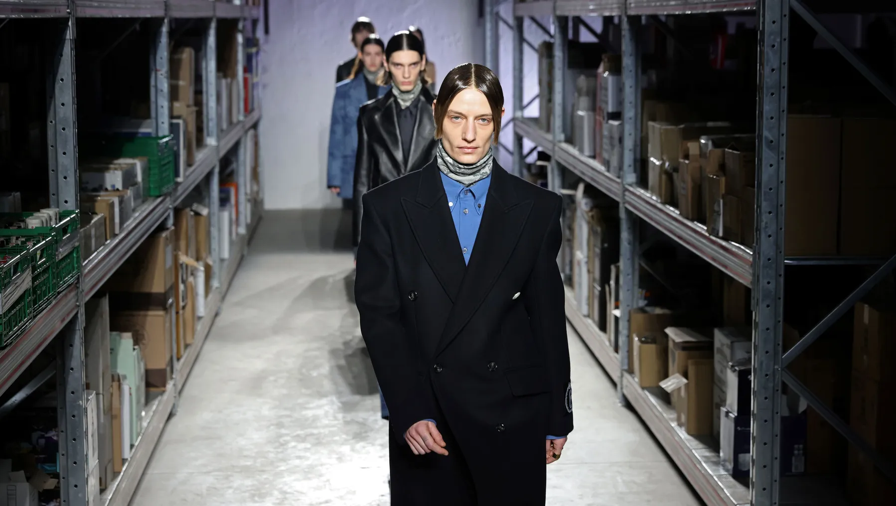 Models walk in a line between metallic warehouse shelves lined with boxes.