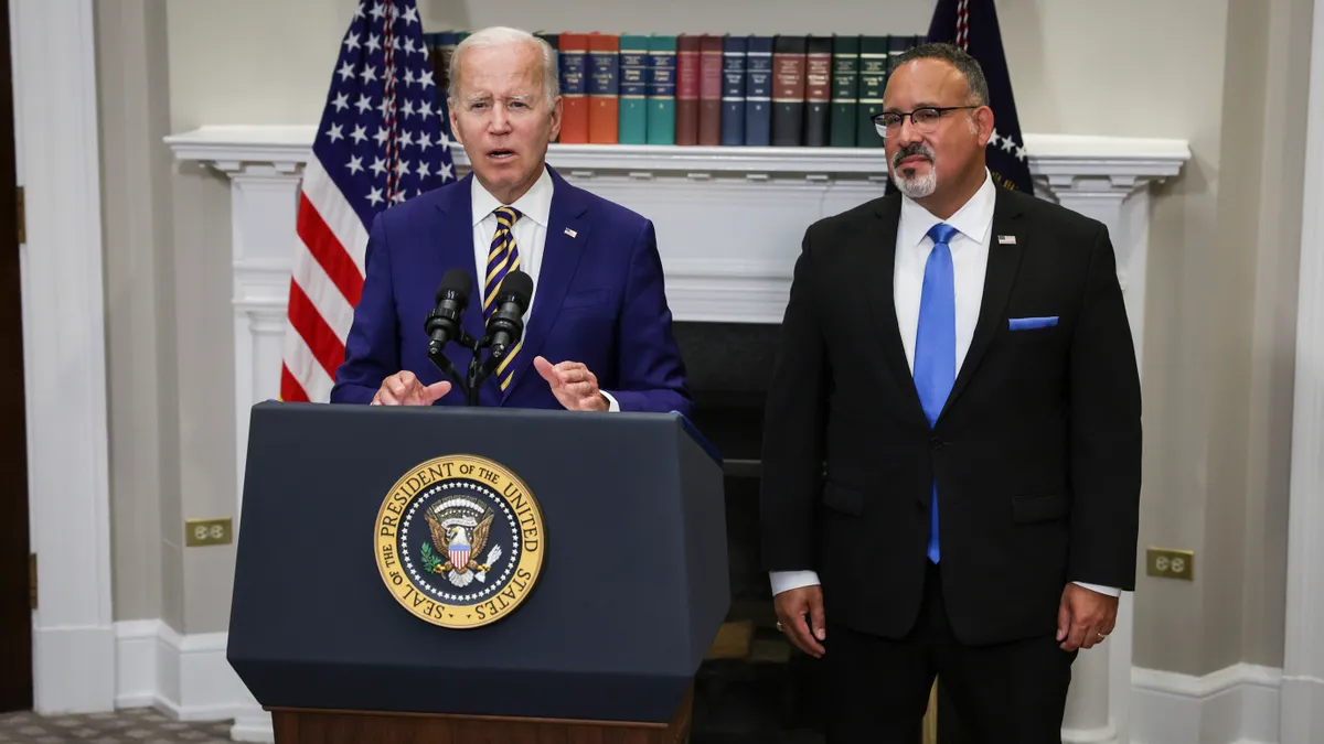 President Joe Biden stands behind a podium while Education Secretary Miguel Cardona stands next to him.