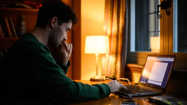 A man in a green shirt sits in a dorm room looking at a laptop screen in lamp light.