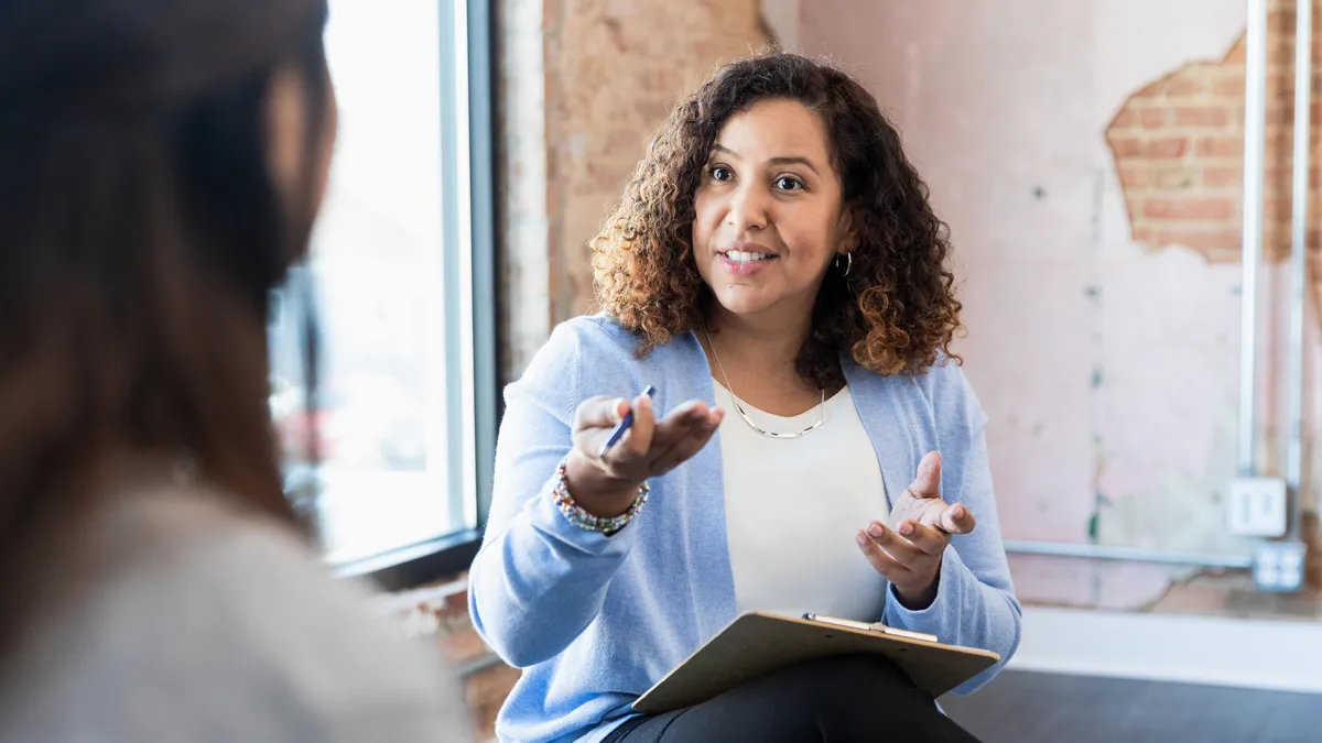 A seated person speaks with another person whose face isn't seen.