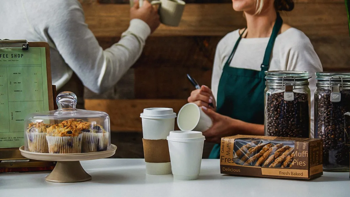 A scene inside a coffee shop with paper cups and food on a counter and a barista using a marker to write on a cup.