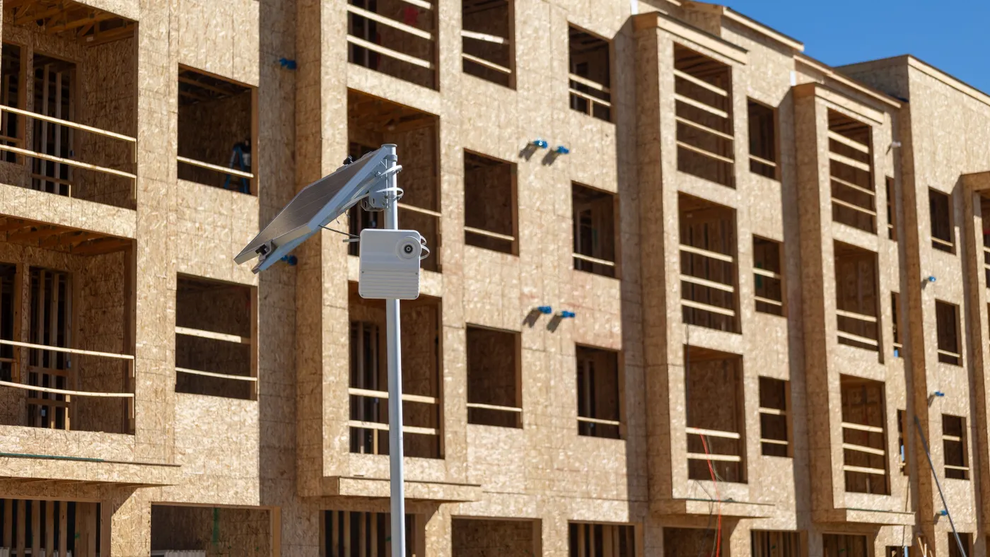 A camera sits on a metal post outside of a brown building on stone amid a bright day.