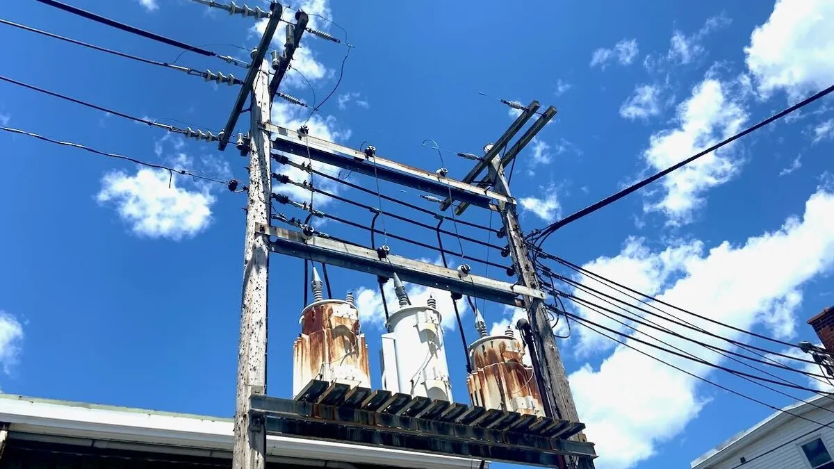 Three electrical transformers sitting on an elevated platform with electric wires crossing in a blue sky overhead.