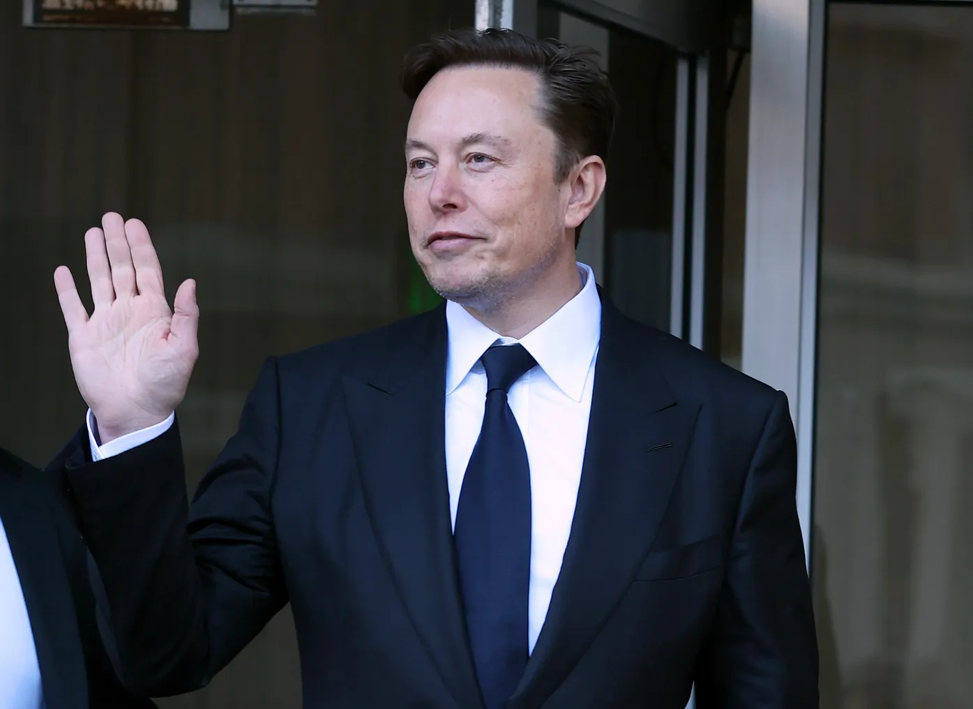 A man in a suit and a navy blue tie waving with his right hand.