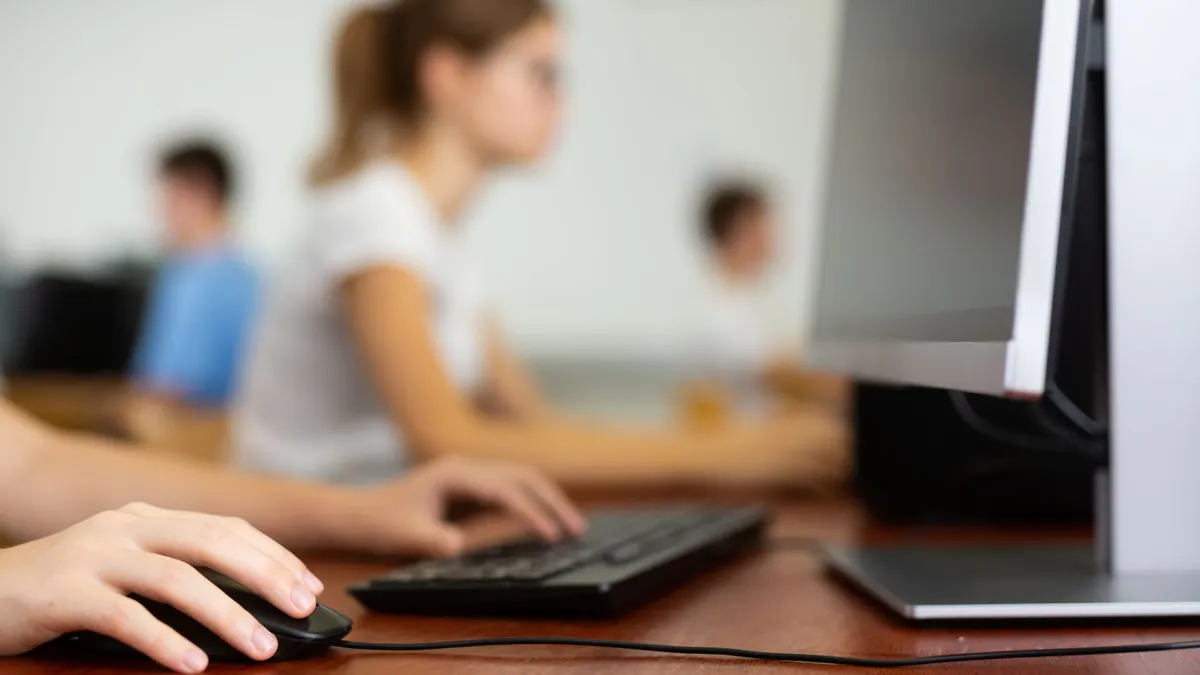 A blurred photo of a teenage student in glasses learning computer science while using a computer in a classroom.