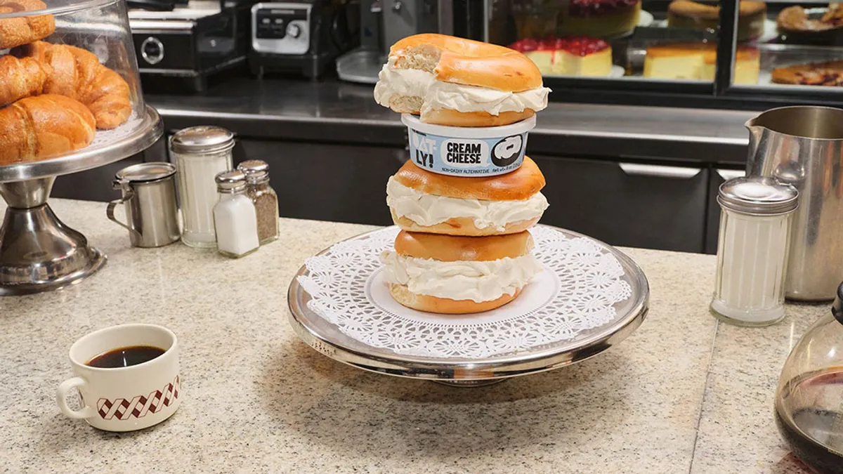 A stack of plain bagels thickly spread with cream cheese, with a container of Oatly Cream Cheese in the pile, sits on a paper doily on a silver platter on a diner counter.