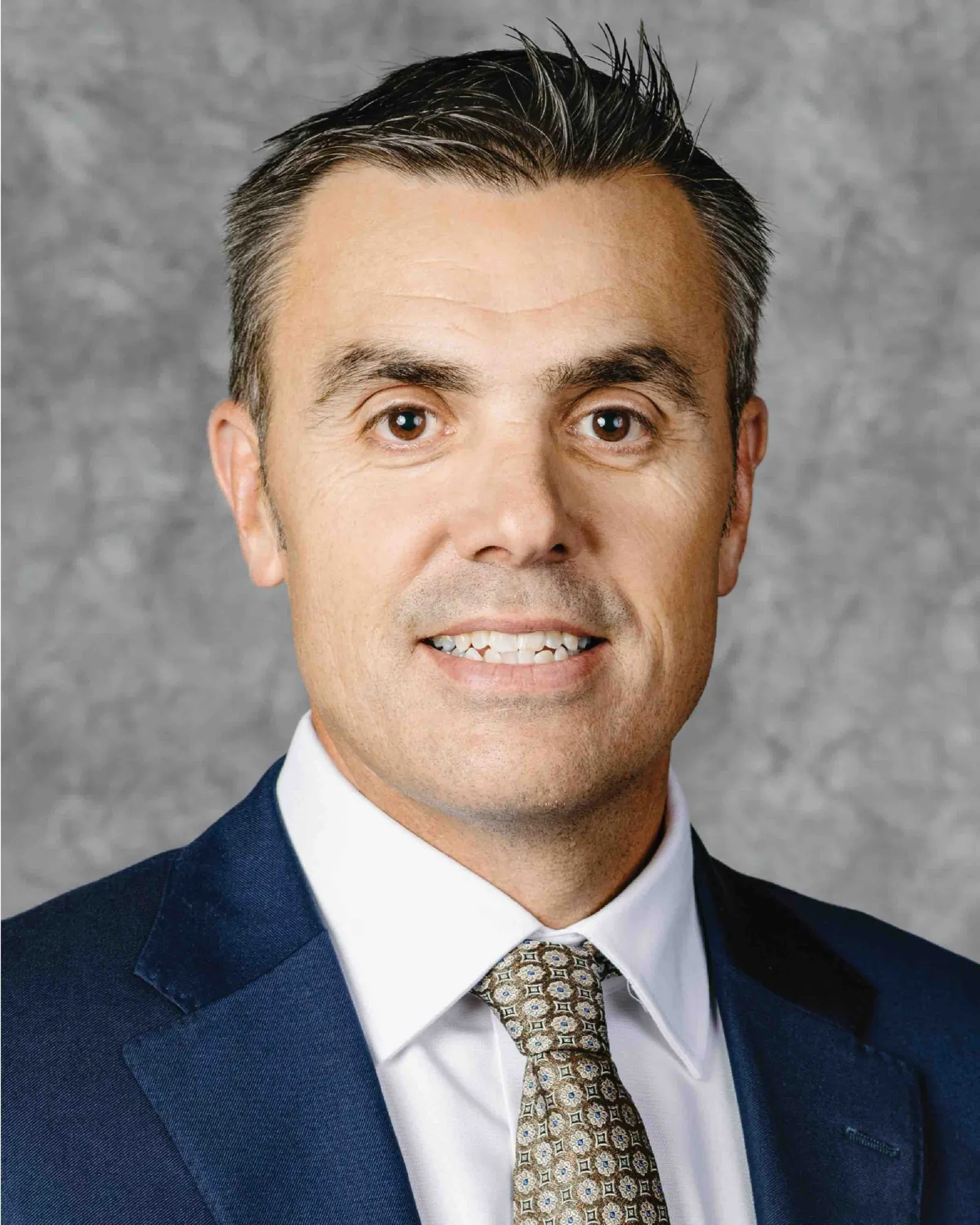 Head and shoulders photo of a man in a blue suit with a brown patterned tie and dark and silver hair.