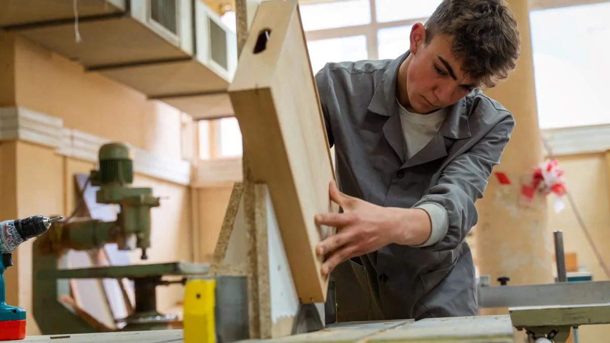 A high school-aged student performs carpentry work in a wood shop.