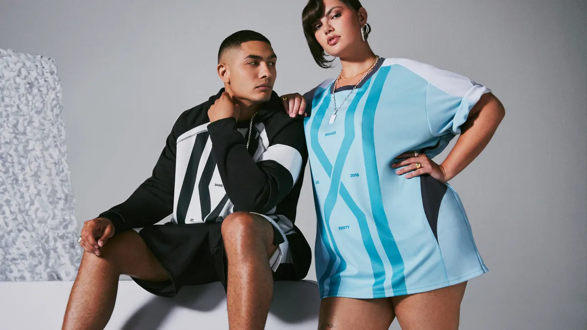 Two models with dark hair pose wearing blue and black Savage X Fenty jerseys.