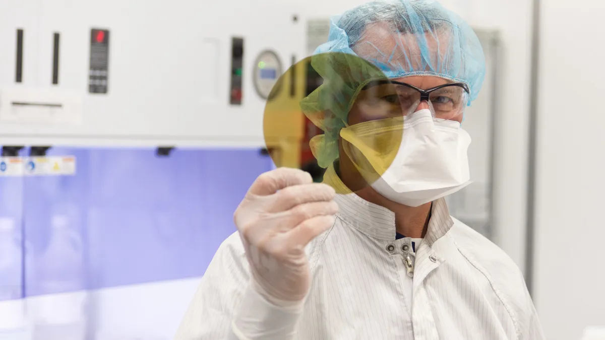 A person with glasses and wearing PPE holds up a yellow wafer in a lab.