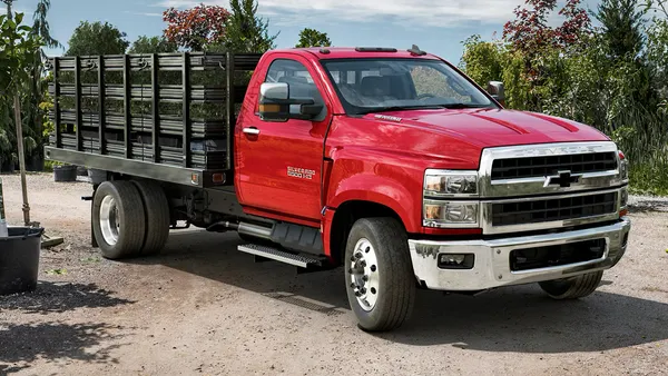 A red 2024 Chevrolet Silverado Chassis Cab parked at a work site with a bridge and construction machinery in the background.