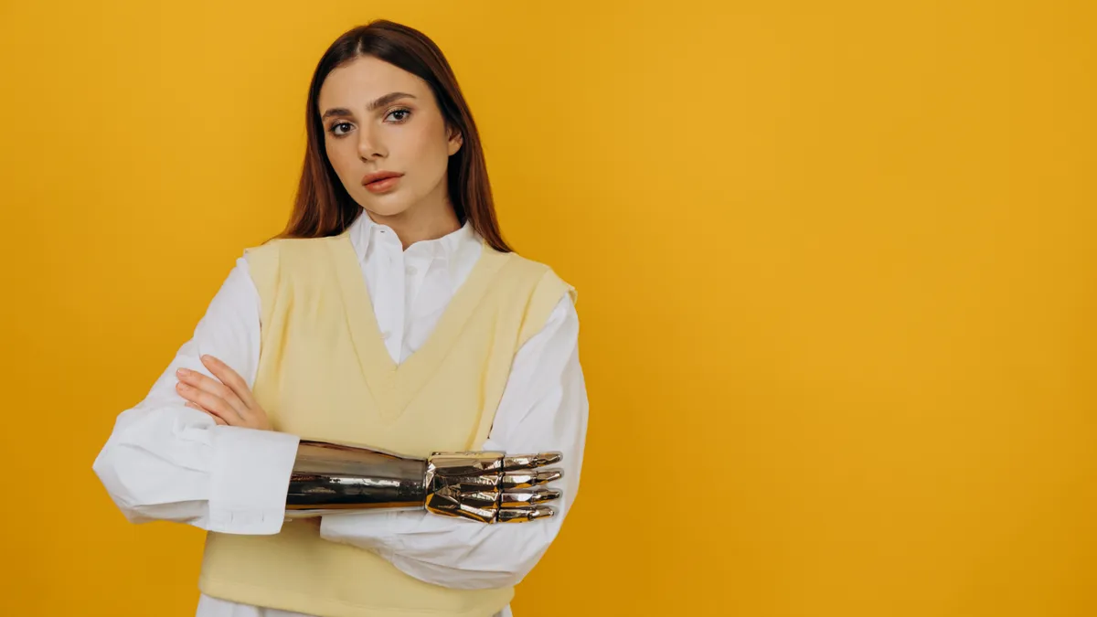 A young White woman with a prosthetic arm wearing a crisp white shirt and a pale yellow sweater vest leans back against a sharp yellow background