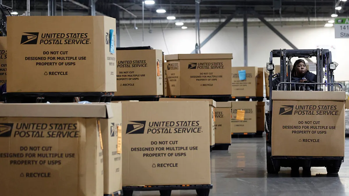 United States Postal Service-branded cardboard boxes stacked in a warehouse, on the right side an employee drives drives a forklift, transporting a box.
