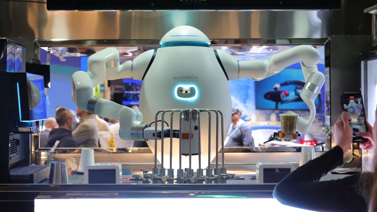 A white oval-shaped robot with arms holds two drinks inside a modern mini kitchen; a person with a phone takes a picture of the robot on the right.