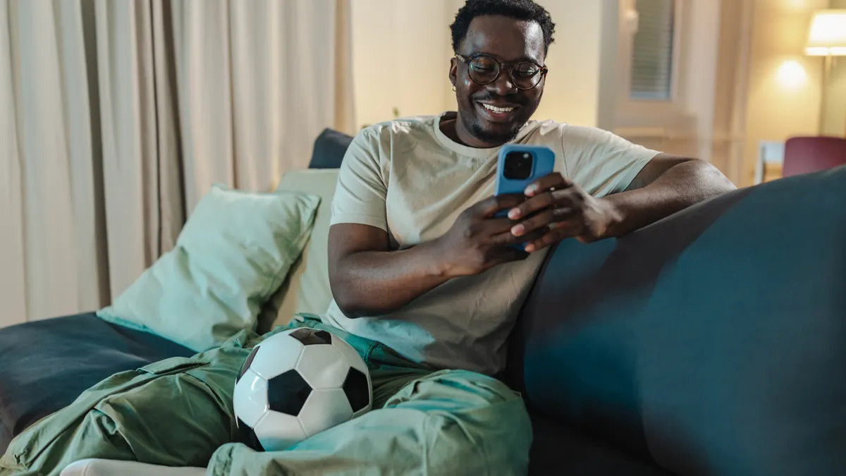 A man with a soccer ball by his feet watches a streaming app on his phone.