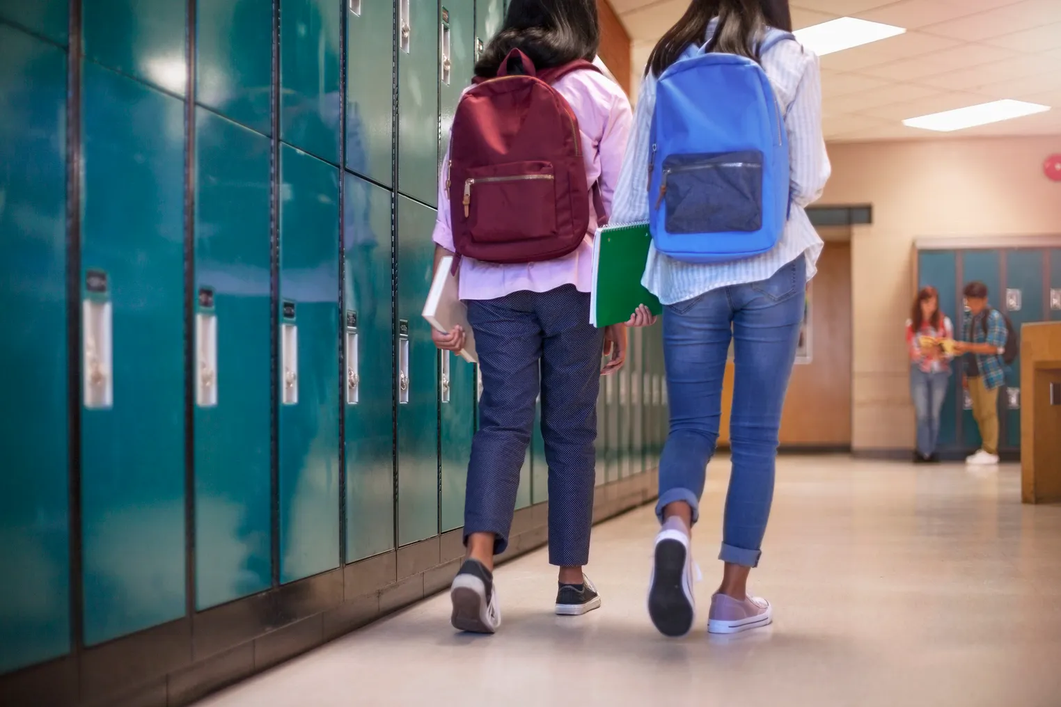 Two middle school students walk down a school hallway next to a row of lockers, backs toward the camera.