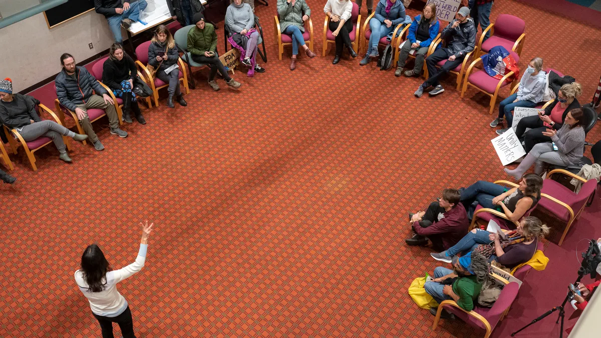 An overhead view shows people sitting in chairs in a U-shape inside a room. One person is standing in front of the people.